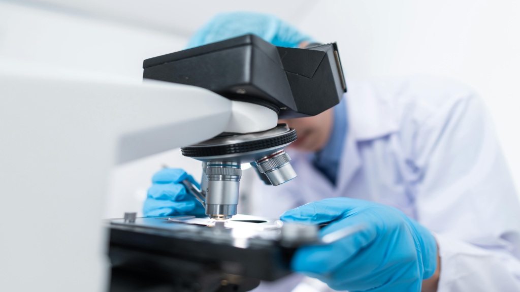 Un científico con bata blanca, guantes y gorro azul observa a través de un microscopio.